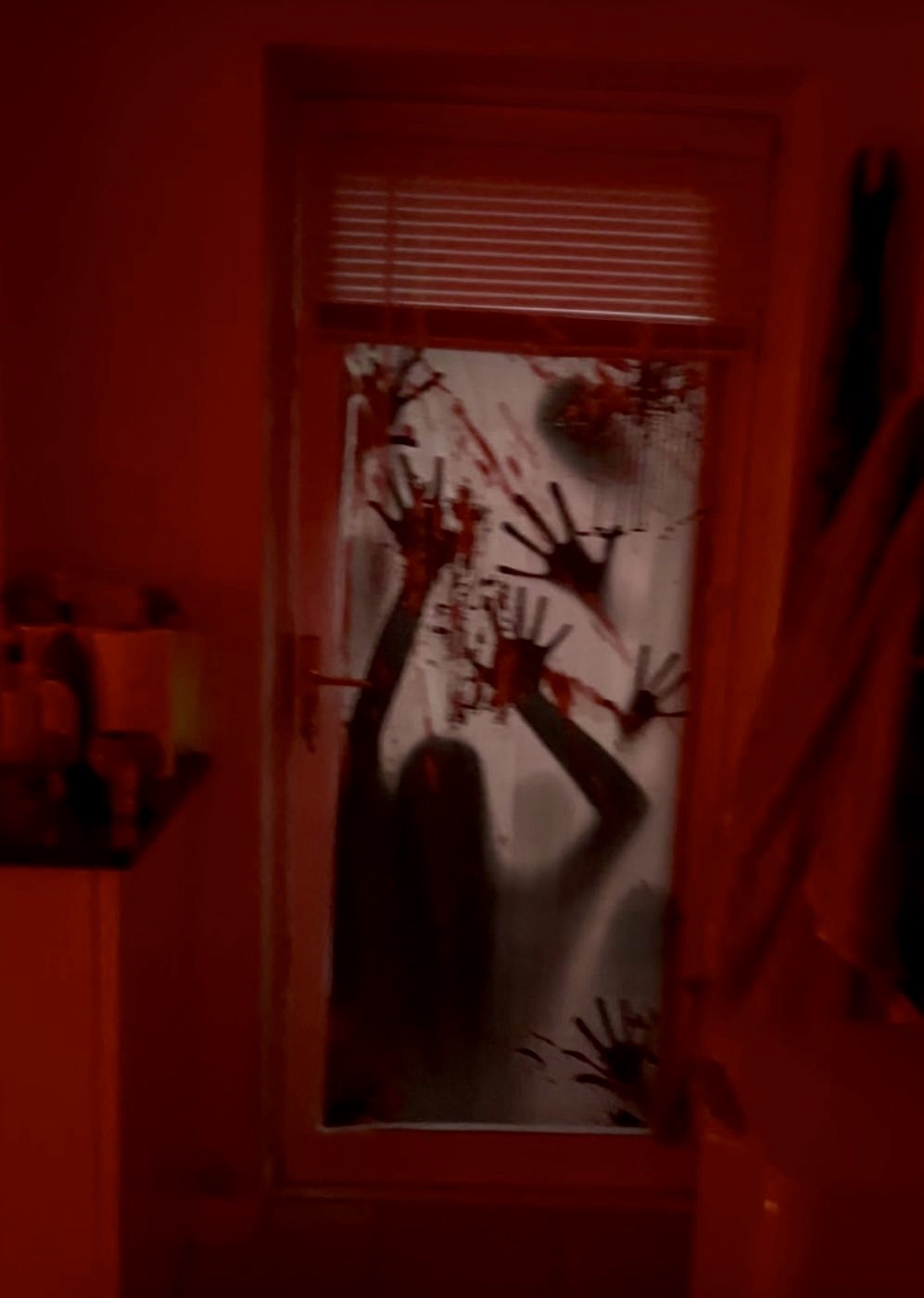 A door covered with a window cling of bloody handprints and a shadowy figure, lit red