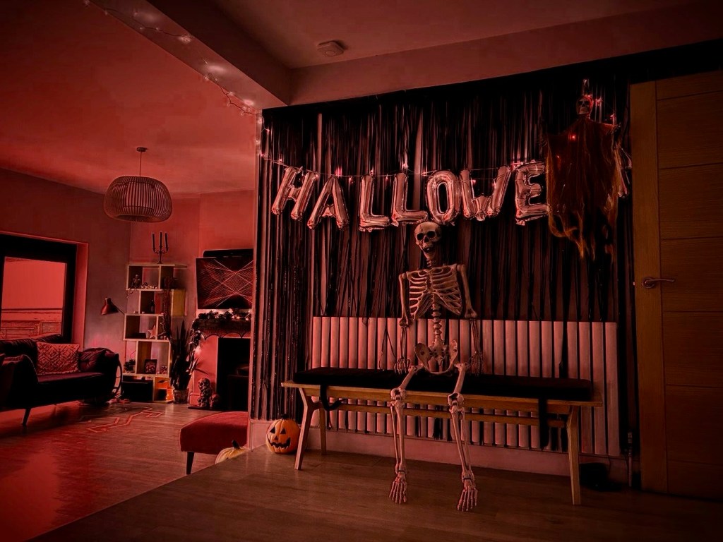 A black tinsel backdrop with silver HALLOWEEN balloon letters and a life-size skeleton sitting on a bench