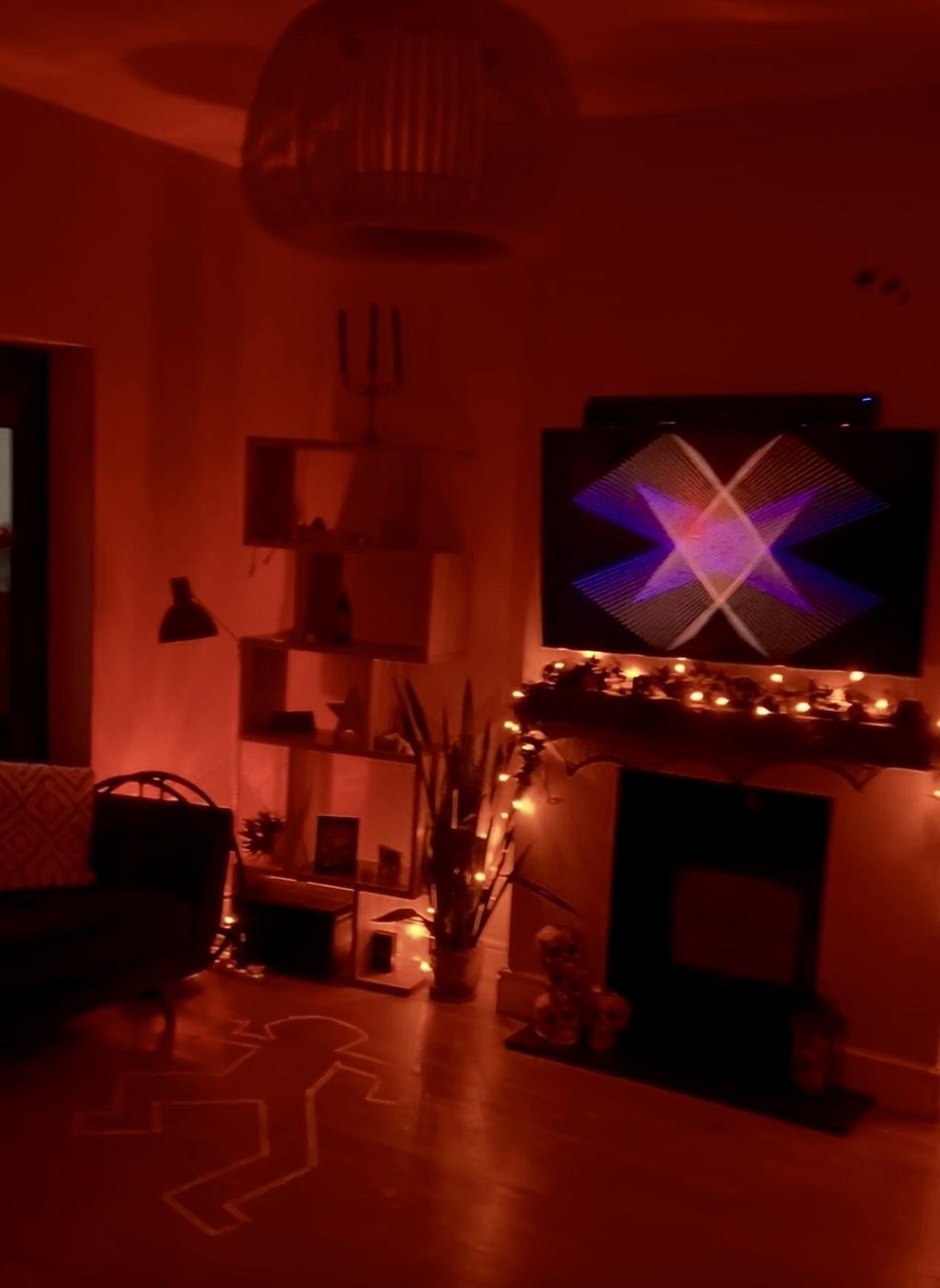 A living room lit red, with fairy lights along the mantelpiece and a body outline taped on the floor